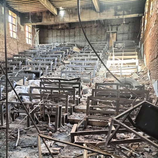 A lecture hall with rows of metal desks and benches heavily damaged, debris and broken wood scattered across the floor, and sunlight entering through windows on the left.